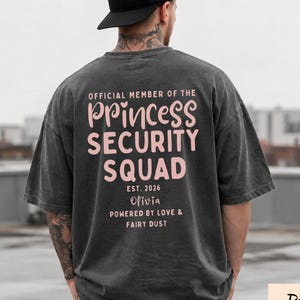 May include: A charcoal gray t-shirt featuring light pink text on the back that reads: OFFICIAL MEMBER OF THE PRINCESS SECURITY SQUAD EST. 2026 Olivia POWERED BY LOVE & FAIRY DUST. The shirt has a loose fit and a vintage-style washed appearance.