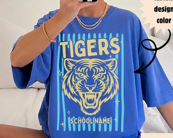 May include: A blue t-shirt featuring a yellow tiger head graphic with the text TIGERS and SCHOOL NAME. The design includes light blue vertical stripes and small star accents. A text box points to the design with the words custom design color.