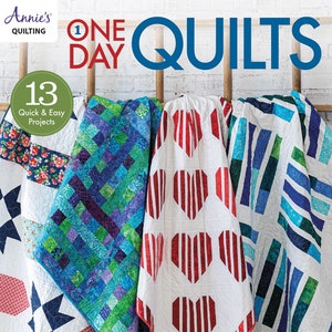 May include: A book cover titled "One Day Quilts" with the Annie's Quilting logo. The cover displays several colorful quilts, including one with red heart shapes and another with blue and green geometric patterns. The book promises 13 quick and easy projects.