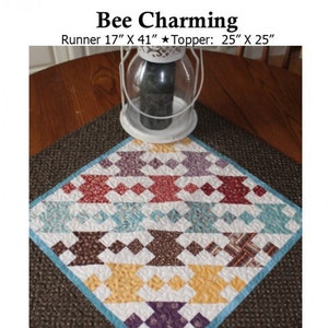 May include: A quilted table runner and topper with a geometric pattern in shades of red, brown, yellow, blue, and purple. The runner is 17 inches by 41 inches and the topper is 25 inches by 25 inches. The text "Bee Charming" and "Creek Side Stitches #192" is visible in the image.