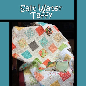 May include: A handmade quilt with a white background and colorful square patches. The quilt features squares in various colors and patterns, including floral and geometric designs. The text "Salt Water Taffy" is at the top.