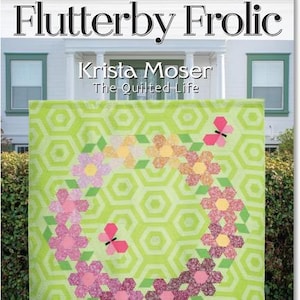 May include: A quilt with a green hexagonal pattern and a floral wreath design. The wreath is composed of pink, purple, and yellow flowers with green leaves and pink butterflies. The text "Flutterby Frolic" and "Krista Moser The Quilted Life" are at the top. The twin size is 76" x 84".