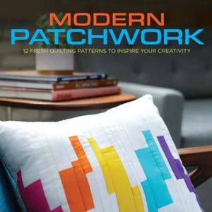 May include: A white pillow with a colorful patchwork design featuring a rainbow of colors including pink, orange, yellow, blue, and purple. The pillow is on a blue couch with a wooden armrest. The text "FAVEQUILTS LEISURE ARTS the art of everyday living" is visible in the bottom right corner of the image.