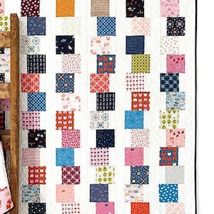 May include: Two quilts made with charm packs. The larger quilt is white with a pattern of colorful squares. The smaller quilt is white with a pattern of colorful squares and stripes. The quilts are displayed on a wooden ladder against a white wall. The text "Charming Stacks" is displayed at the bottom of the image.