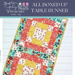 May include: A quilted table runner with a floral pattern in red, yellow, and teal. The runner is rectangular, with a design that includes squares and rectangles. The text "ALL BOXED UP TABLE RUNNER" is at the top.