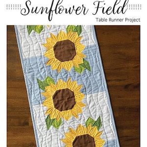 May include: A quilted table runner with a sunflower design. The runner features three large sunflowers with yellow petals and brown centres, set against a light blue and white background. The finished size is 36.8 cm x 105.4 cm. The text "Sunflower Field" is at the top.
