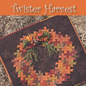 May include: A quilt pattern called "Twister Harvest" designed by Becky Cogan for Needle Love. The quilt features a wreath design with autumn colors and a brown background. The quilt is displayed on a bed of straw with pumpkins and greenery in the foreground.