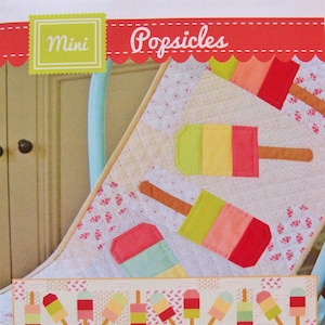 May include: A colorful quilt featuring popsicle designs in shades of pink, green, and yellow. The word "Popsicles" is written in a playful font. The quilt is on a light blue chair.