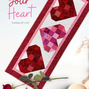 May include: A quilted table runner with three heart shapes in shades of red and pink. The runner is 16 inches by 42 inches and features the text "Follow Your Heart". The design includes a white background and a red dotted border.