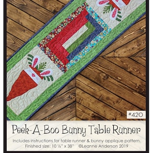 May include: A table runner pattern featuring a bunny peeking out of a carrot. The pattern is called "Peek-A-Boo Bunny Table Runner" and is part of "The Whole Country Caboodle". The finished size is 10 1/2 inches by 38 inches.