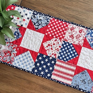 May include: A patriotic table runner featuring a patchwork design with red, white, and blue fabrics. The runner showcases various patterns, including stars, gingham, floral prints, and stripes, with a quilted texture and a star-studded border.