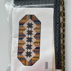 May include: A packaged quilting kit featuring a colorful star-patterned design. The kit includes fabric rolls and a pattern that measures 18 x 42 inches. The fabrics have geometric and plaid patterns in shades of blue, yellow, and gray.