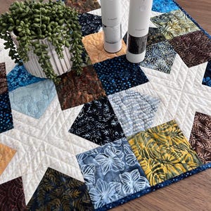 May include: A quilted table runner featuring a star pattern in white fabric surrounded by squares of various batik prints in blue, brown, yellow, and black. A white ribbed pot with a trailing green plant and two white salt and pepper mills.