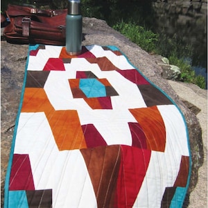 May include: A colorful patchwork quilt with geometric shapes in shades of brown, red, orange, and teal. The quilt is laid out on a large rock with a silver thermos nearby.