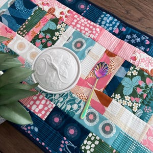 May include: A rectangular fabric table runner featuring a patchwork design of various patterns in shades of pink, teal, navy blue, and white, including strawberry and floral motifs. A white lidded ceramic jar and a rainbow-colored shell spoon.