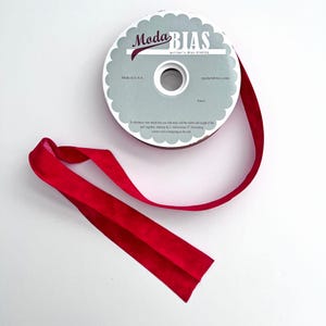 May include: A spool of Moda Bias quilter's bias binding tape in a solid deep red color. The round cardboard label features the text Moda BIAS, quilter's Bias binding, Made in U.S.A., and modafabrics.com, with a scalloped edge design.
