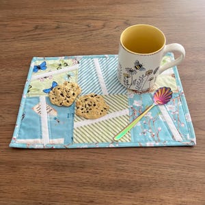 May include: A patchwork placemat with a blue and white floral and striped pattern, holding a white mug with a yellow interior and floral design, two chocolate chip cookies, and a rainbow spoon. The placemat has a stitched edge.