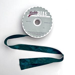 May include: A spool of Moda Bias quilter's bias binding with a scalloped edge label. The spool features the text Moda BIAS and includes a length of dark teal fabric tape with a mottled, textured pattern extending from the center.