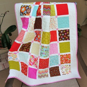 May include: A patchwork quilt with a white background and squares of various colors and patterns, including pink, red, green, brown, blue, and yellow. The quilt is edged with a pink and white trim.