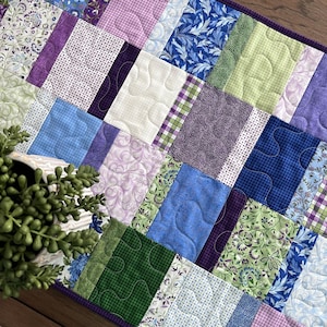May include: A rectangular quilted table runner featuring a patchwork design of various fabric patterns in shades of blue, green, purple, and white. The quilt is stitched with a continuous meandering pattern and finished with a dark purple border.
