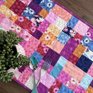 May include: A rectangular quilted table runner featuring a patchwork design of various floral and geometric fabric squares in shades of pink, purple, orange, blue, and white. The fabric squares are stitched together with a purple border.