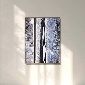May include: A framed aerial photograph of a winter landscape. The image features a road and a river winding through snow-covered trees, with shades of white, gray, and black. The artwork is displayed on a neutral-colored wall.