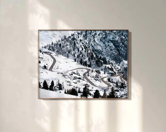 May include: A framed photograph of a winding mountain road in a snowy landscape. The road curves through the snow-covered terrain, with evergreen trees dotting the slopes. The sky is overcast, and the overall tone is cool and serene.