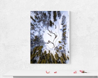 May include: An aerial view of a winter landscape featuring a snow-covered area surrounded by evergreen trees. A winding river cuts through the snow, and a small white building is visible. The image is framed in a white border.