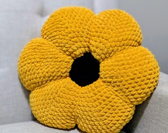 May include: A plush, yellow flower-shaped pillow with a black center. The pillow is made of a textured, soft material and is resting on a gray surface. The flower has large, rounded petals.