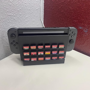 以下が含まれることがあります： 黒いニンテンドースイッチ本体と、24本のゲームカートリッジを収納した黒いスタンド。本体は白い表面の上に置かれている。