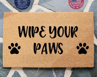 May include: A brown coir doormat with the text "Wipe Your Paws" in black lettering. Two black paw prints are on either side of the text.