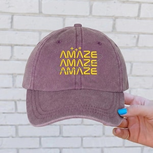 Puede incluir: Gorra de béisbol malva con la palabra "AMAZE" bordada en amarillo. La palabra se repite tres veces, con un diseño de planeta estilizado alrededor de la palabra del medio. La gorra tiene una visera curva y una corona redondeada.