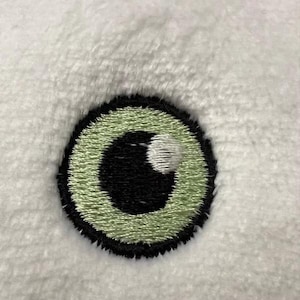 Peut inclure: Gros plan sur l'œil d'un jouet en peluche, brodé avec un iris vert et une pupille noire.