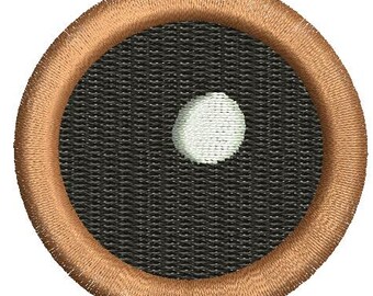 May include: Embroidered patch featuring a black circle with a single white dot, surrounded by a brown border. The patch has a textured appearance, suggesting it is made of fabric or thread. The design is simple and graphic.