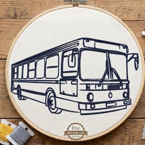 May include: A circular wooden embroidery hoop containing white fabric with a dark blue stitched outline of a city bus. The design features a detailed bus silhouette. Surrounding the hoop are gold-toned scissors, spools of blue, yellow, and gray thread, and several small fabric swatches in various shades of blue.