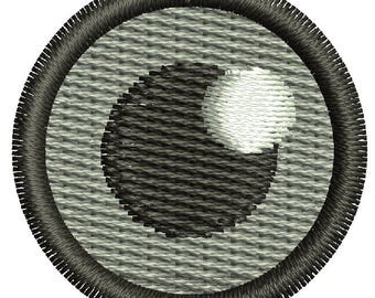 May include: A black and white embroidered eye patch with a gray background. The eye patch is circular and has a stitched border.