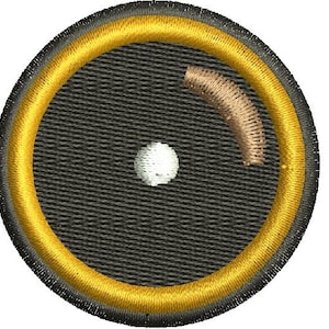 May include: Embroidered patch with a circular design. The patch features a black background with a gold border and a white dot in the center. A curved, tan-colored element is also present.