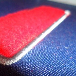 Op de afbeelding: Een close-up van een rode stoffen patch die op een blauwe stoffen oppervlak is genaaid. De patch heeft een witte rand en is gestikt met een decoratieve rand.