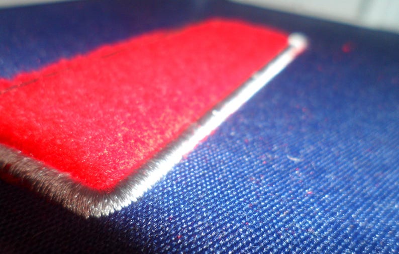 Op de afbeelding: Een close-up van een rode stoffen patch die op een blauwe stoffen oppervlak is genaaid. De patch heeft een witte rand en is gestikt met een decoratieve rand.