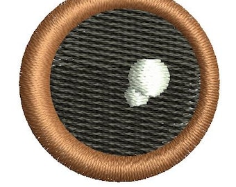 May include: Embroidered patch with a brown border and a black and gray textured background. A white, abstract shape is in the center. The patch is circular.