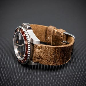 May include: A stainless steel wristwatch with a black dial and a red and black bezel. The watch has a brown leather strap. The watch is set against a dark background.