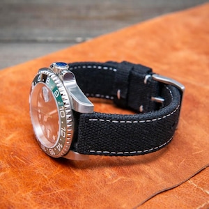 Puede incluir: Una correa de reloj de lona negra con costuras blancas, mostrada sobre una superficie de cuero marrón. La correa está diseñada para un reloj con una esfera redonda y un bisel plateado.