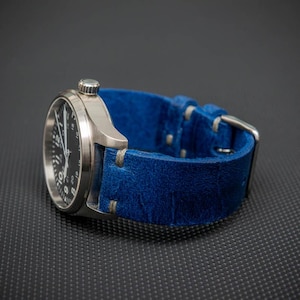 Könnte beinhalten: Eine Armbanduhr mit silbernem Gehäuse und schwarzem Zifferblatt, kombiniert mit einem leuchtend blauen Lederarmband. Die Uhr hat ein klassisches Design mit sichtbarer Krone und Schnallenverschluss. Das Armband ist etwa 2,5 cm breit.