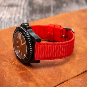 Puede incluir: Un reloj negro con una correa de nylon roja. El reloj tiene una esfera negra con marcas blancas y un bisel plateado.