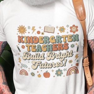May include: White t-shirt with the text "KINDERGARTEN TEACHERS Build Bright Futures!" in a retro font. The design includes a sun, flowers, a book, and a rainbow. The shirt is worn by a person.