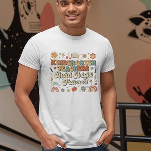 May include: White t-shirt with a colorful graphic that reads "Kindergarten Teachers Build Bright Futures!" The design includes a sun, book, pencils, flowers, and rainbows. The shirt is worn by a person in blue jeans.