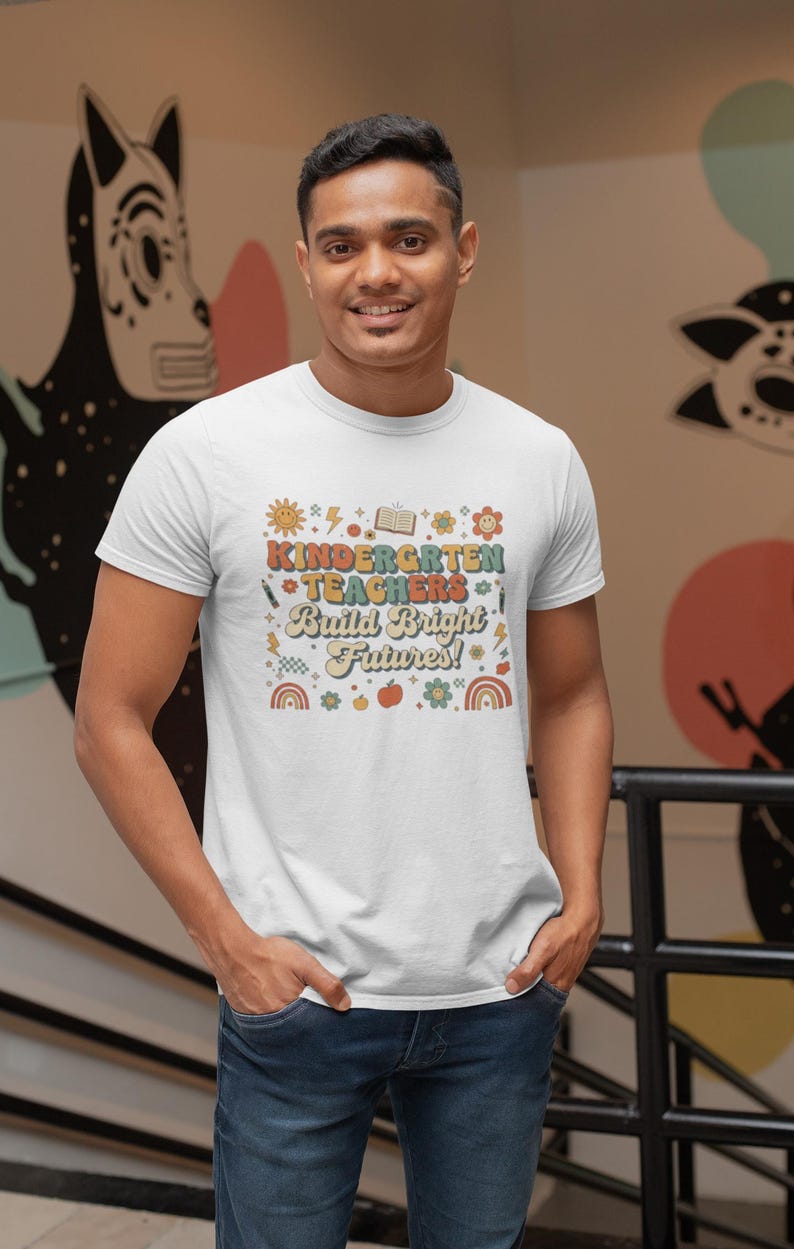 May include: White t-shirt with a colorful graphic that reads "Kindergarten Teachers Build Bright Futures!" The design includes a sun, book, pencils, flowers, and rainbows. The shirt is worn by a person in blue jeans.