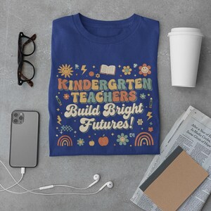 May include: A navy blue t-shirt with the words "KINDERGARTEN TEACHERS Build Bright Futures!" in a retro font. The design includes a sun, flowers, rainbows, and an apple. Other items include glasses, a phone, and a coffee cup.