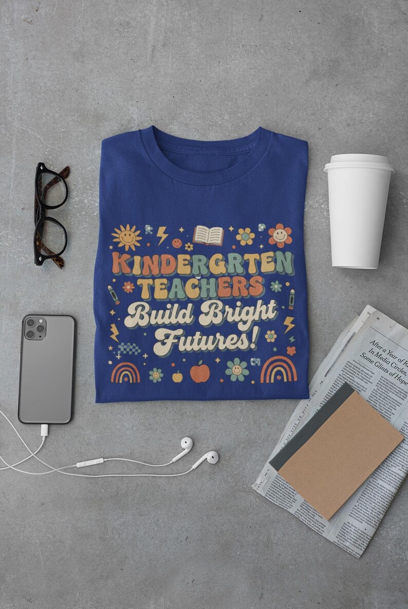 May include: A navy blue t-shirt with the words "KINDERGARTEN TEACHERS Build Bright Futures!" in a retro font. The design includes a sun, flowers, rainbows, and an apple. Other items include glasses, a phone, and a coffee cup.