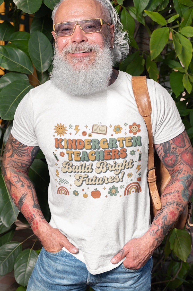 May include: White t-shirt with the text "KINDERGARTEN TEACHERS Build Bright Futures!" in a retro font. The design includes a sun, flowers, a book, and a rainbow. The shirt is worn by a person.
